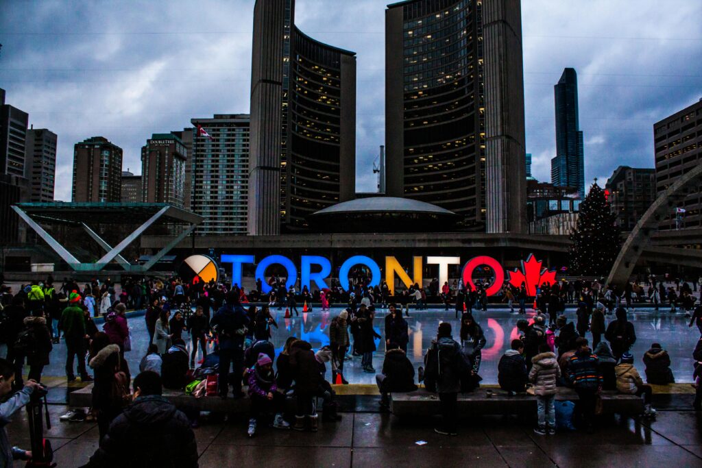 Toronto sign at Nathan Phillips Square with people skating and gathering in front of city buildings under cloudy skies – symbolizing Canada’s welcoming culture for immigrants and students.