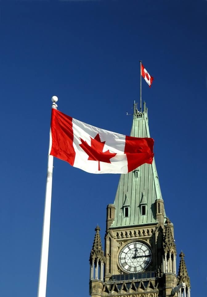 Canadian flag flying proudly in front of Parliament Hill with the iconic clock tower in Ottawa under a clear blue sky.